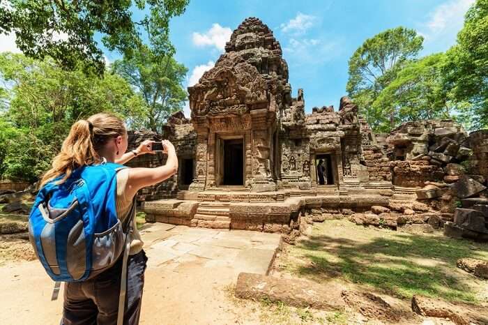 Young female tourist with smartphone taking picture of the gopura under blue sky near the entrance to ancient Preah Khan temple in Angkor