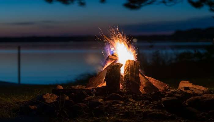 The view of a bonfire on the side of a lake while camping.